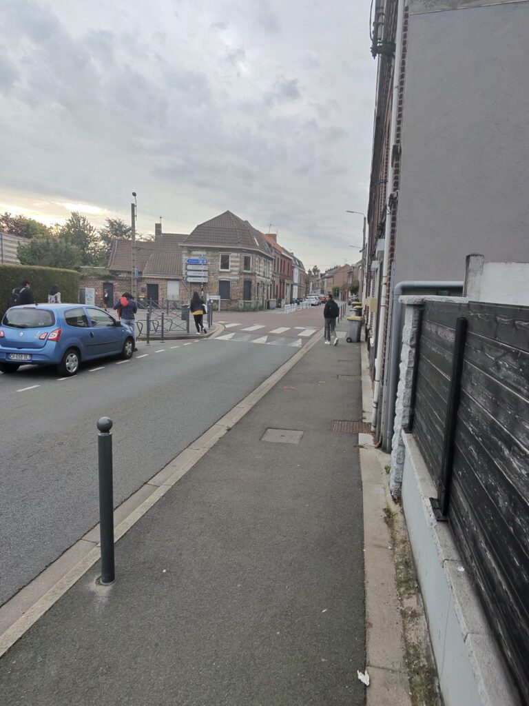 Trottoir à Ronchin avec un potelet sur le cheminement, du mobilier urbain et des poubelles plus loin, illustrant les enjeux d’accessibilité de l’espace public.