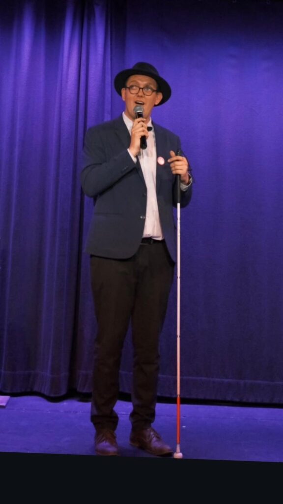 Nicolas Karasiewicz sur la scène du Spotlight à Lille, devant un rideau violet, portant un chapeau noir et une veste bleue, un micro à la main et sa canne blanche devant lui.