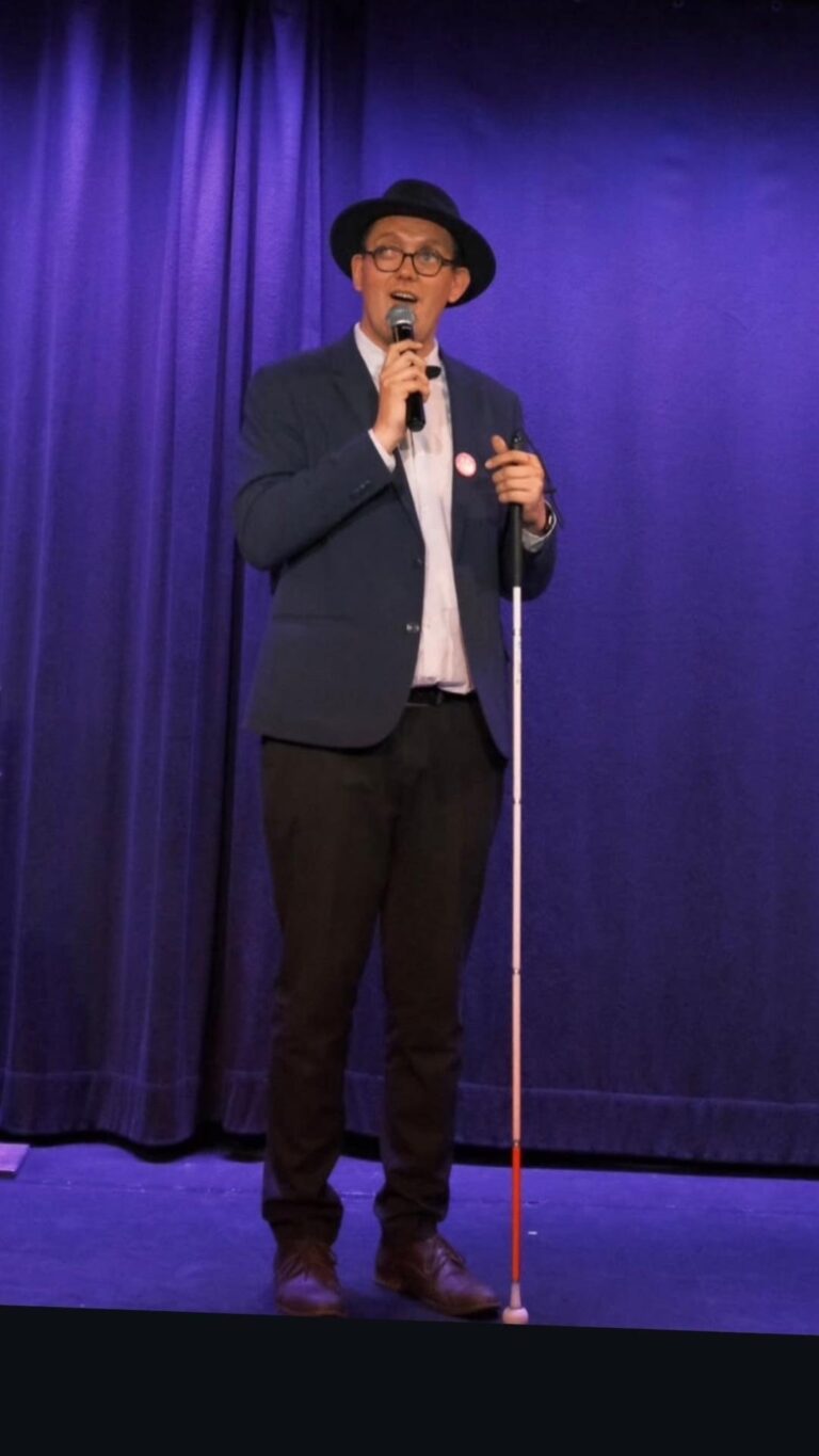 Nicolas Karasiewicz sur la scène du Spotlight à Lille, devant un rideau violet, portant un chapeau noir et une veste bleue, un micro à la main et sa canne blanche devant lui.