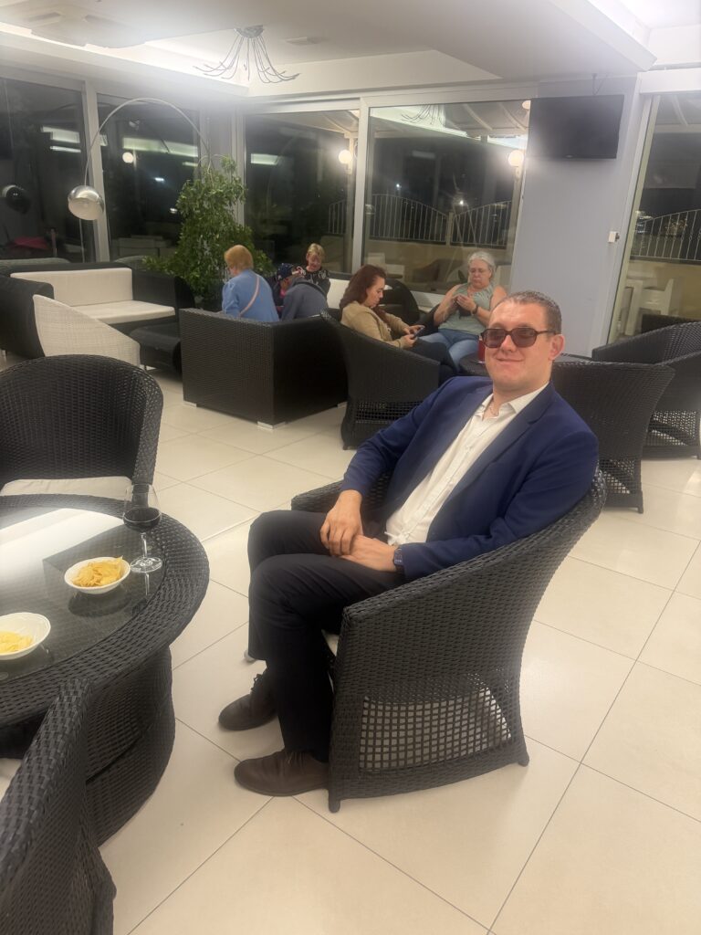 Nicolas, en veste bleue et chemise blanche, sourit assis dans un fauteuil en rotin noir au bar de son hôtel à Jesolo. Un verre et des coupelles sont posés sur la table devant lui.